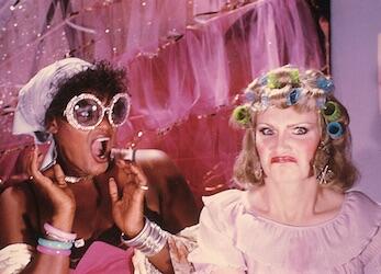Film still of two women in extravagant pink costumes in front of pink set, surprised and disgusted