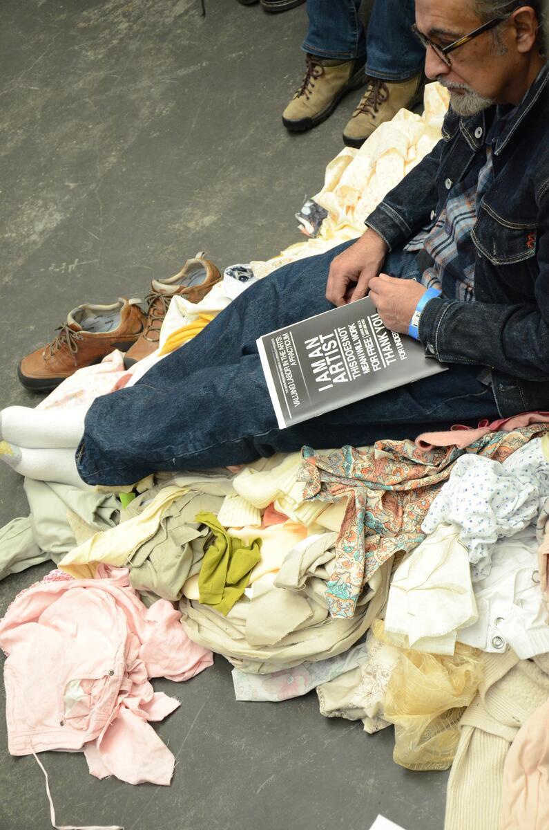 Man sitting on pile of garments, shoes off, with magazine in his lap