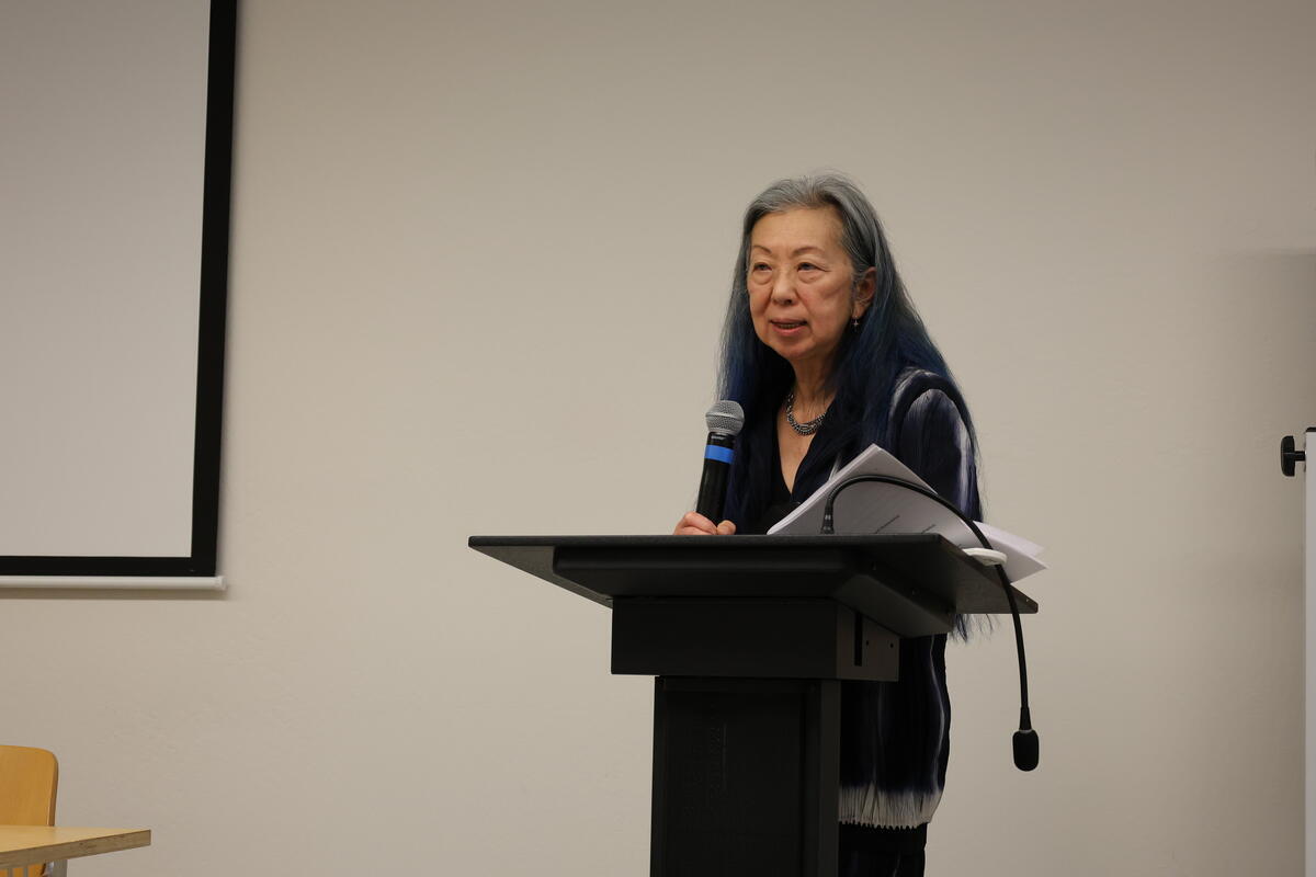 Person with long blue hair speaking at a podium holding a microphone and papers.