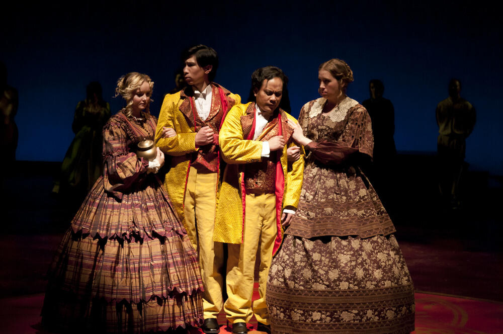 Four actors in period costumes on a dimly lit stage, with two men in yellow tailcoats and two women in long dresses, one in a plaid dress and the other in a dark brown dress.