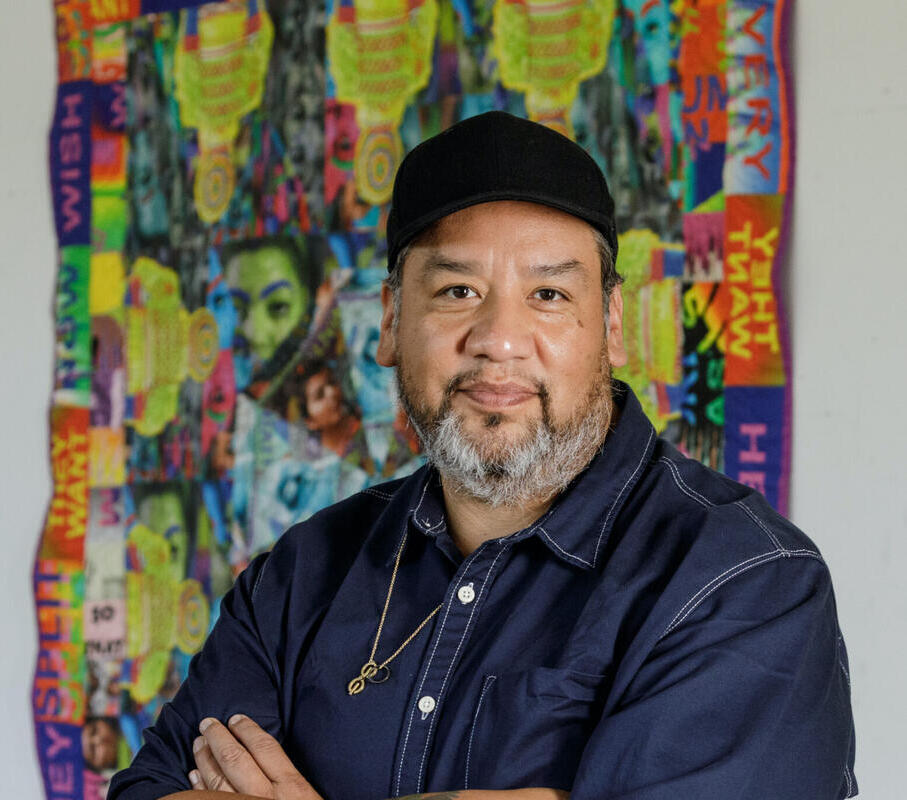Portrait of Jeffery Gibson wearing a denim shirt and a black cap in front of a colorful artwork