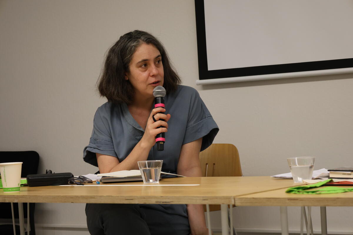 Person speaking into a microphone while seated at a table with papers and a glass of water.