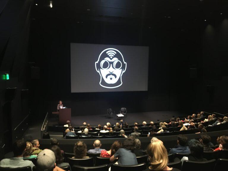 Theater with image of man in pilot goggles with mustache and wifi signal on this helmet projected