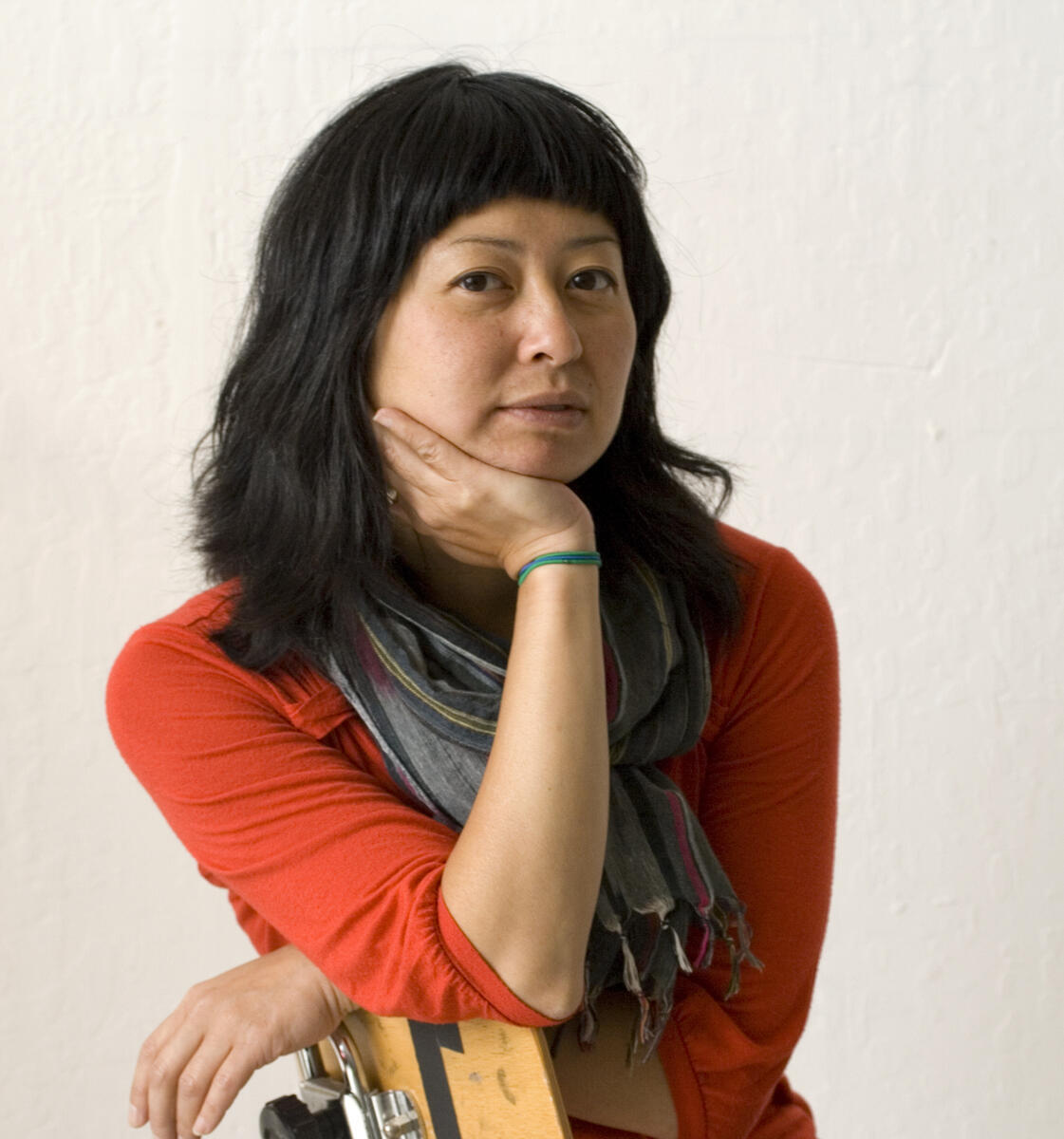 Portrait of a woman with black hair, wearing a grey scarf and red-orange top, resting her face on her hand.