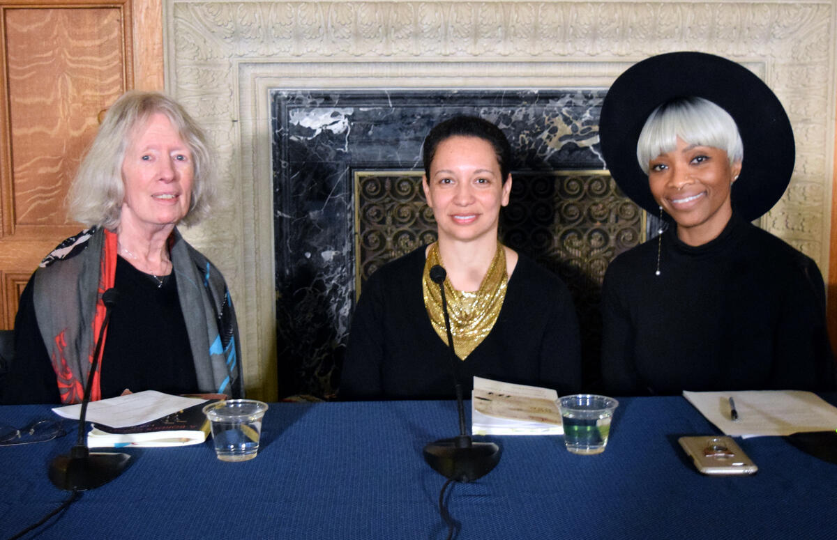 Three people sitting at a table with microphones, in front of a decorative wall. Two have books and one has a phone on the table.