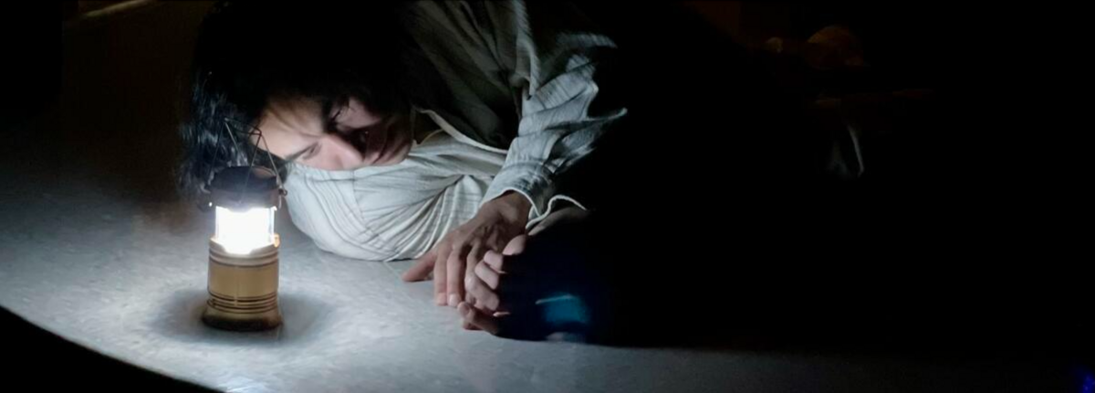 Person lying on the floor next to a small bright lantern in a dark room.