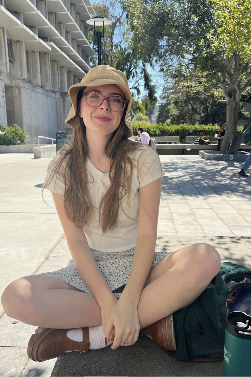 Eva Whitney Smiling woman with glasses and a sunhat sitting cross legged outside