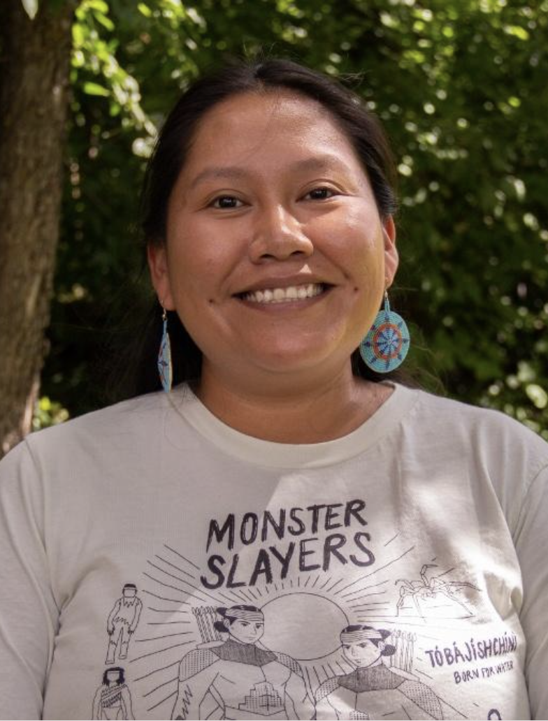 Blossom Johnson wears a white T-shirt with a "MONSTER SLAYERS" illustration and text standing in front of lush green foliage. She is wearing with large, patterned circular earrings.