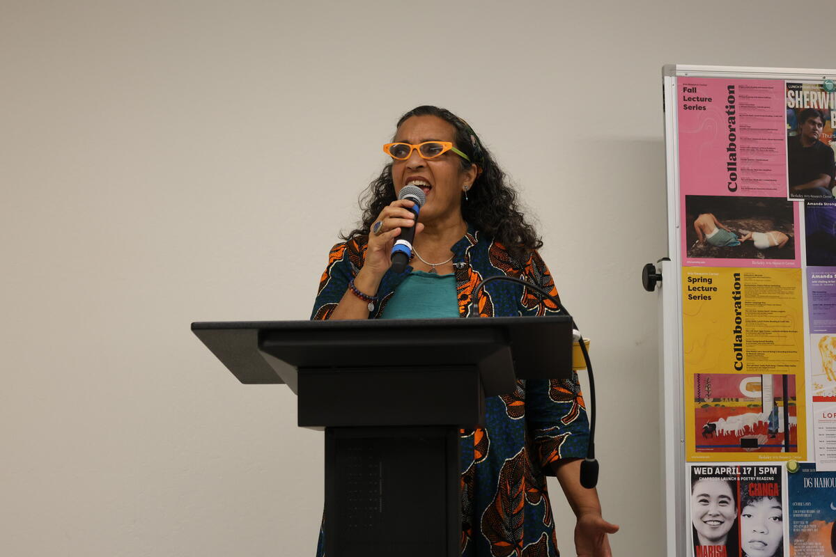 Person speaking into a microphone at a podium with event posters in the background.