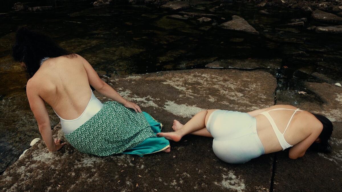 Two women in swimsuits sitting on rocks by a river, their backs facing us. They wear swimsuits and underwear.