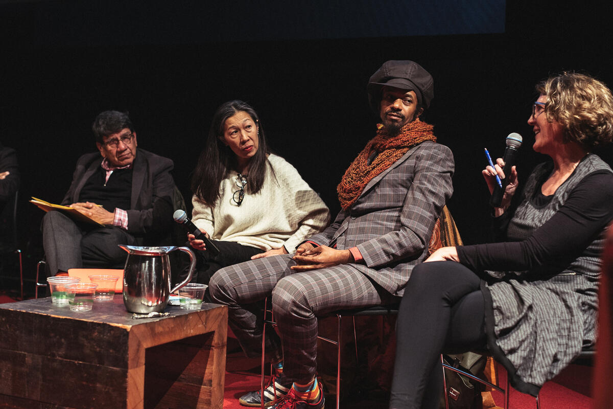 A panel discussion with four people on stage, featuring microphones and a table with a silver pitcher and cups.