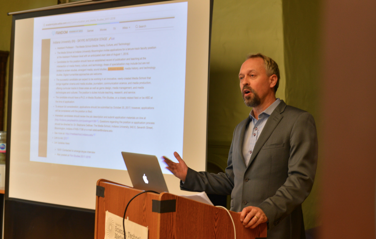 Man in a gray suit speaking at a podium with a laptop and a presentation projected behind him.