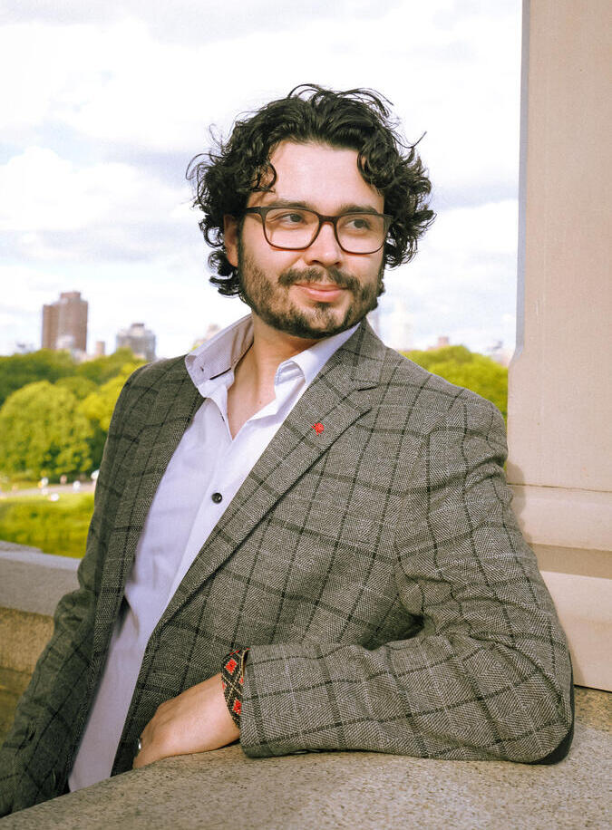 man in plaid jacket wearing glasses leaning against a wall with skyline behind him