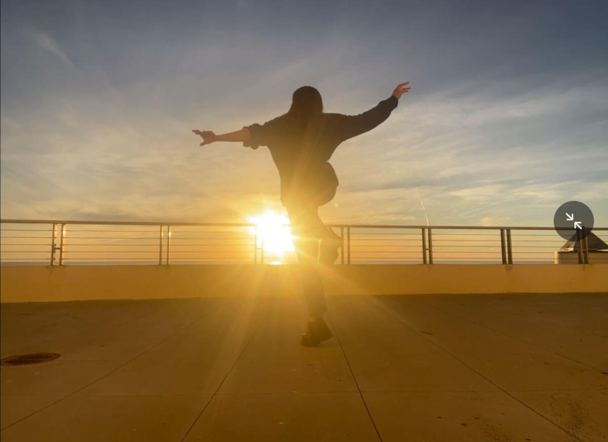 A silhouette of a jumping with arms outstretched, facing the sun as it sets on the horizon. The sun's rays are shining through the railing, casting long shadows on the ground.