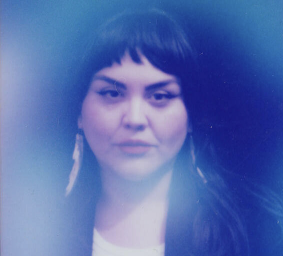 A person with dark hair and earrings enveloped in a blue haze.