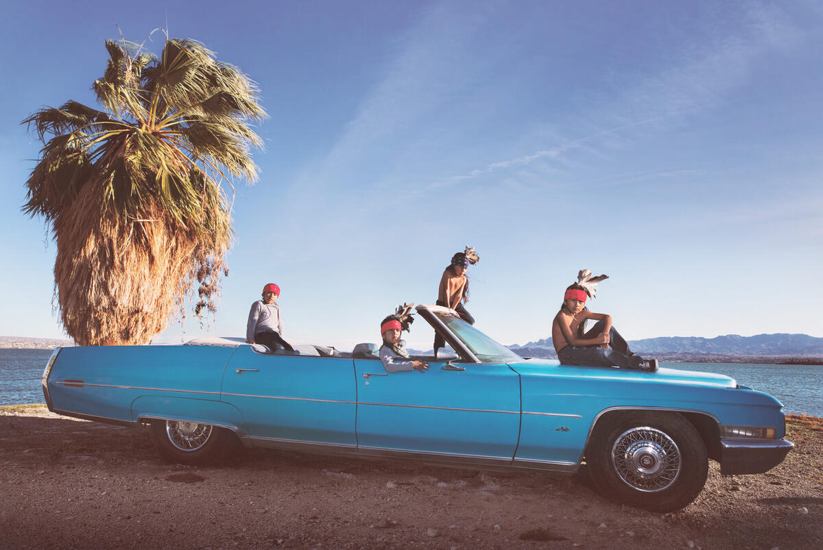 Four boys in Native headdresses with feathers sit in a blue vintage car near lake with a palm tree
