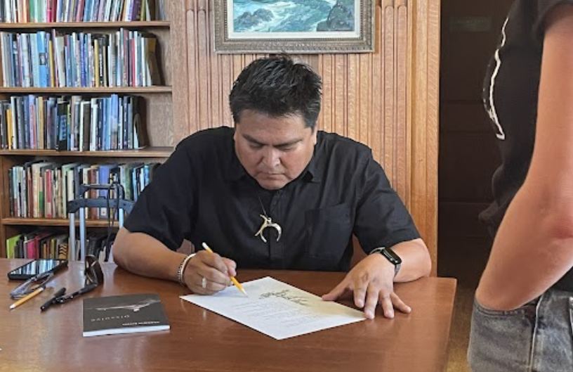 Person writing at a desk with books, pens, and a book titled "Dissolve" while a person stands nearby. Behind is a bookshelf labeled "Poetry" and a painting of ocean waves.