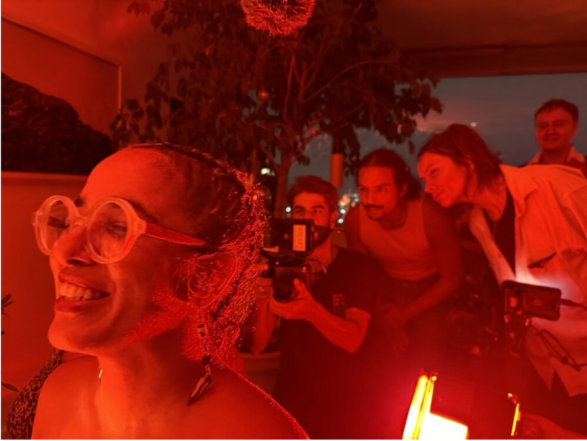 Person smiling with large glasses in a red-lit room, three people in the background focusing on a camera.