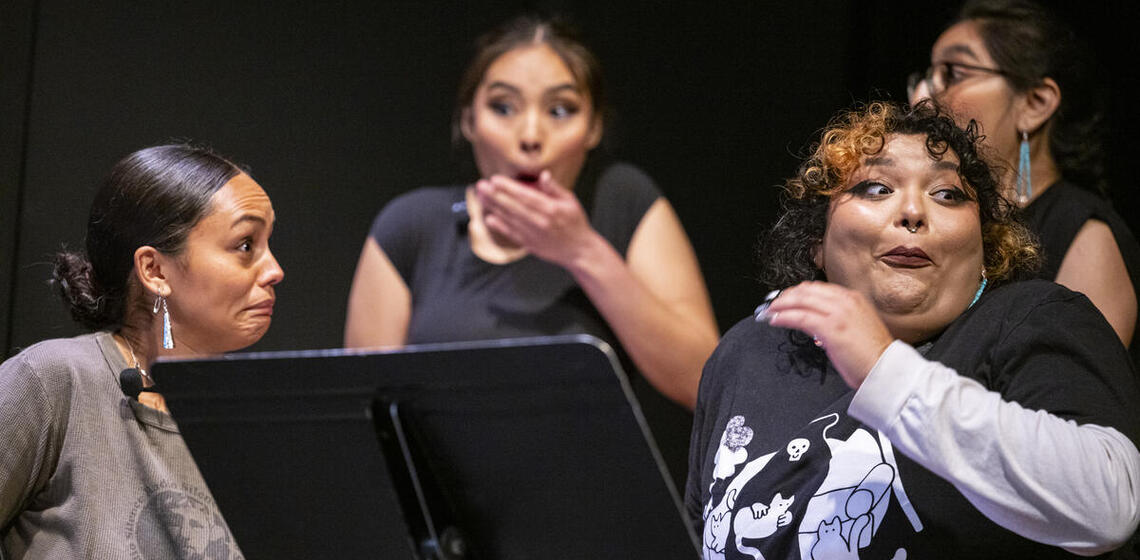 Three women on a stage display dramatic expressions and gestures during a performance, with expressions of surprise and involvem