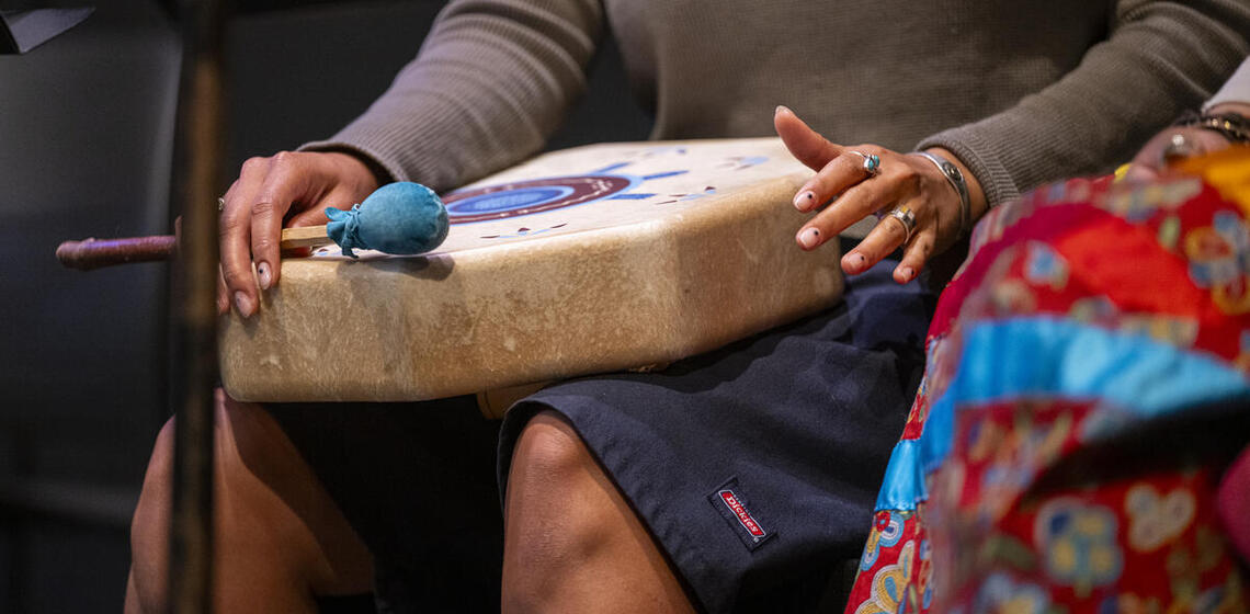 Person playing a hand drum placed on their lap, with a drum mallet on top and colorful fabric visible.