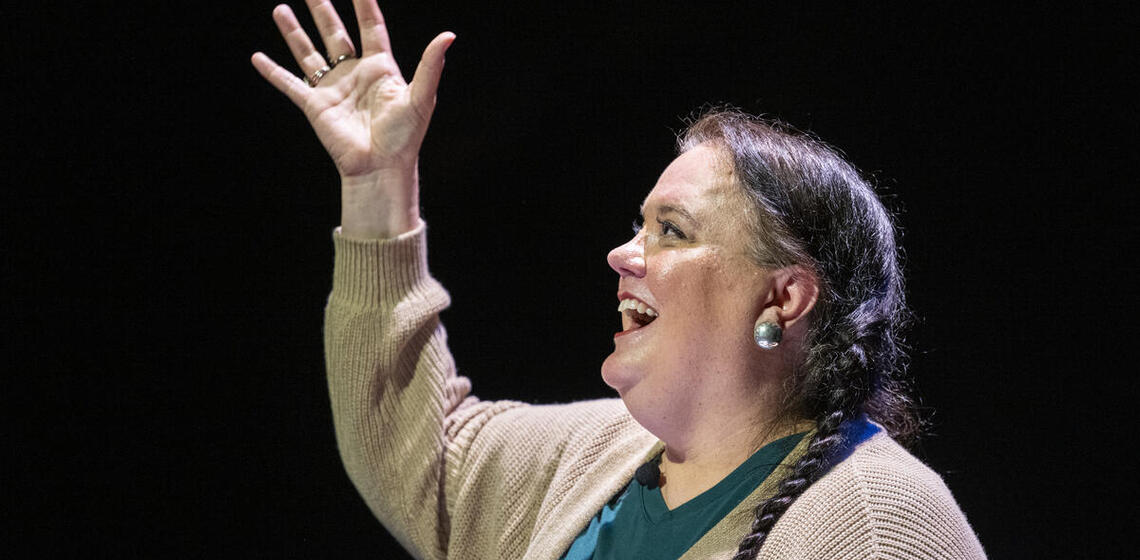 A woman with a braid over her shoulder smiles and gestures while presenting on stage, holding a black notebook. There is no text