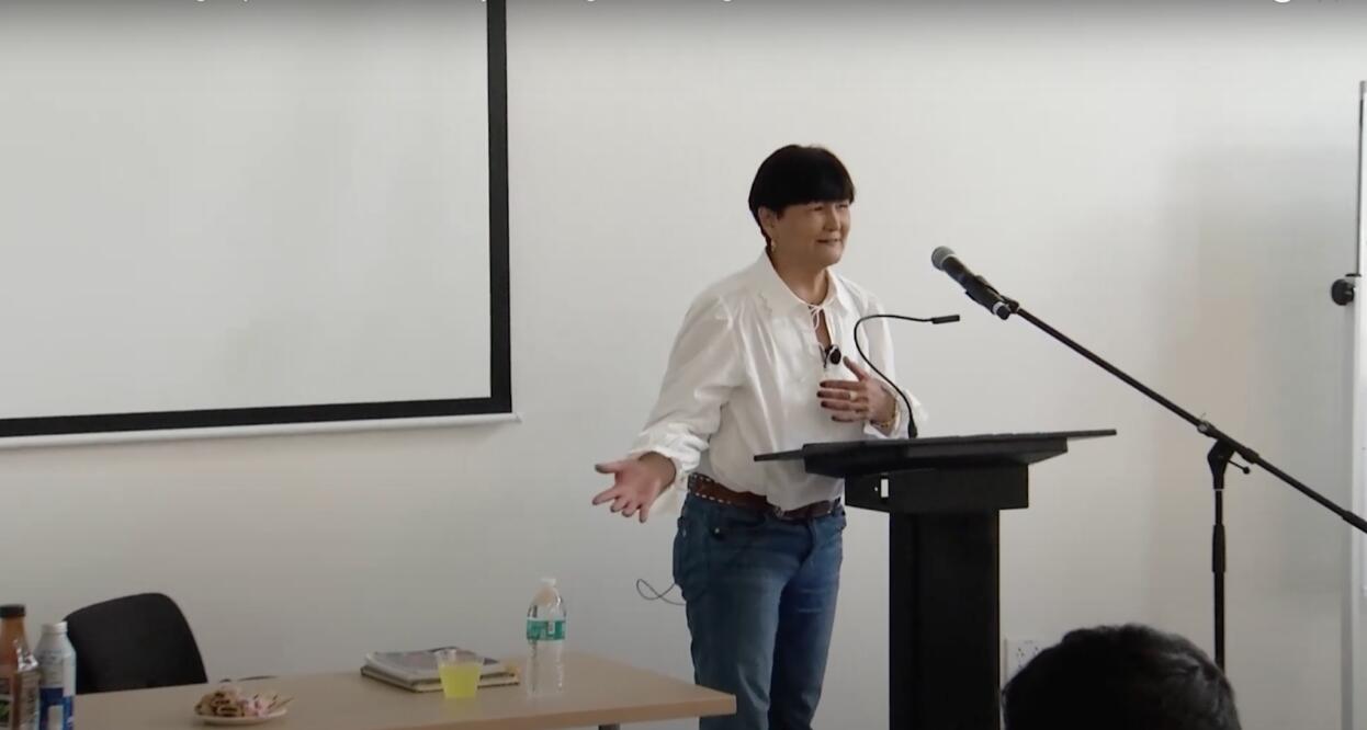 Speaker at podium gestures while talking with a microphone stand next to them. A table with various items is nearby.