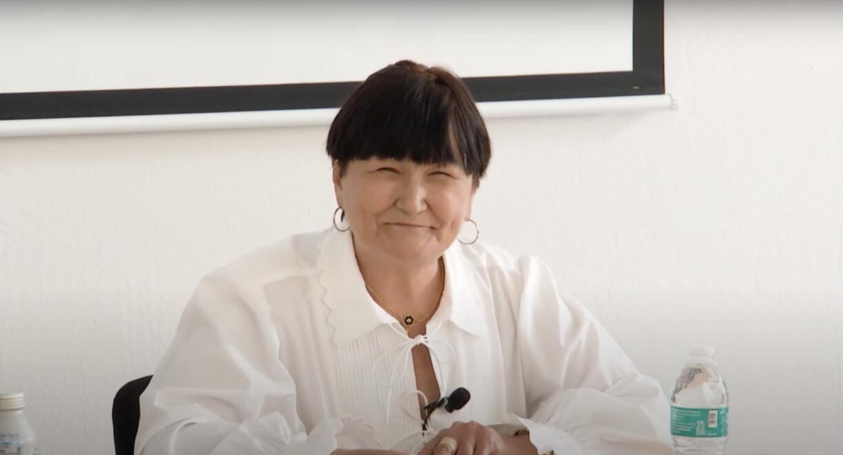 Person with short, dark hair smiling while seated at a table, wearing a white shirt with a scalloped collar and hoop earrings. A water bottle is on the table.