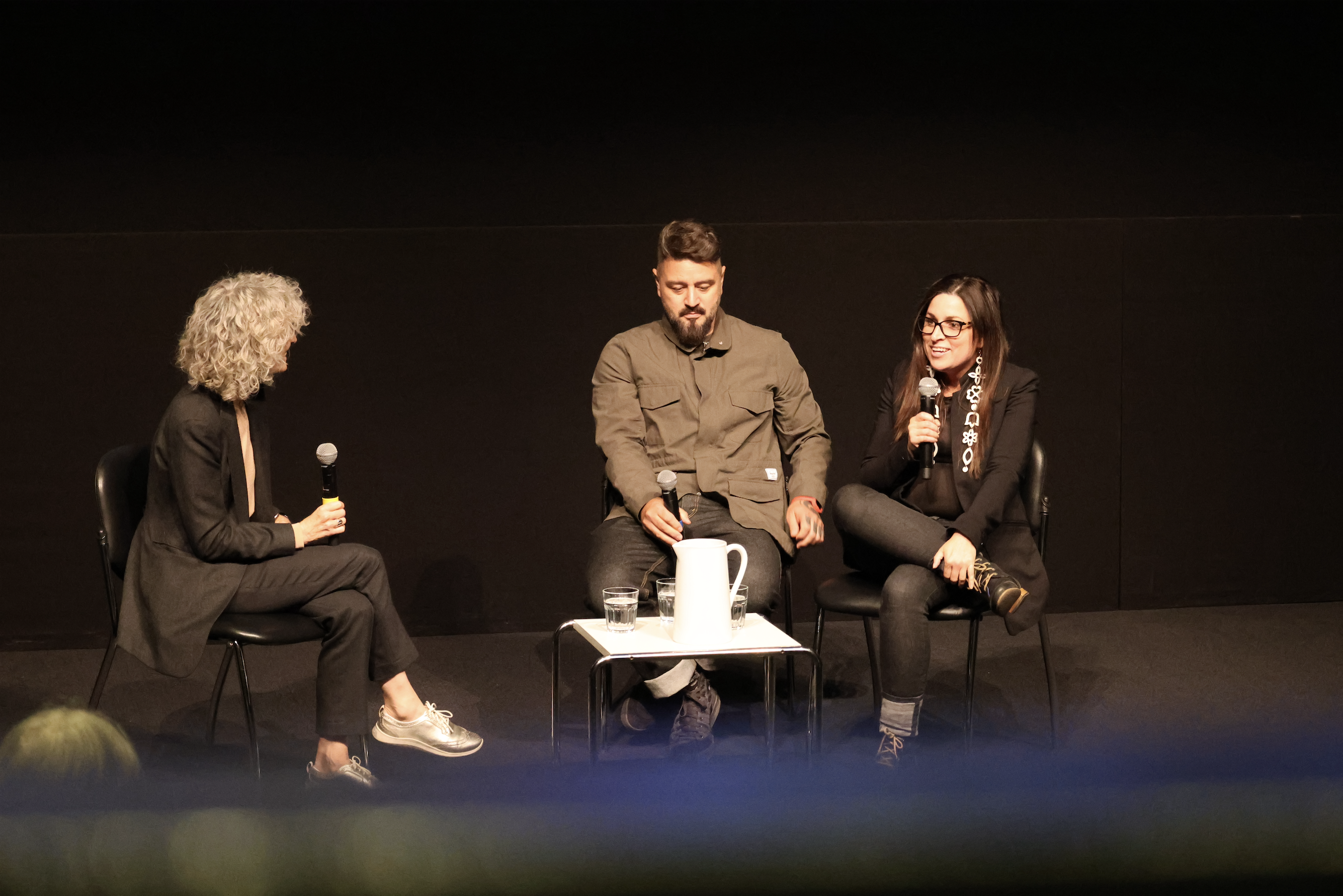 Three individuals are seated in a panel discussion. The woman on the left holds a microphone. The man and woman on the right also hold microphones, smiling and engaged. A small table with a water pitcher and glasses is in front of them, set against a blac