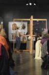 Person in overalls presenting artifacts to a seated audience in an art gallery.