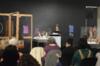 Woman giving a lecture at a podium in a dimly lit room, with an audience seated in front of her and display cases in the background.