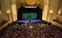 Grand theater interior with occupied red seating, orchestra in front of the stage, and ornate golden curtains.