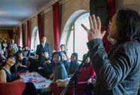 Man speaking to an audience in a room with arched windows and red curtains.