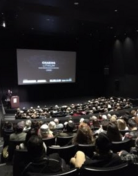 A full auditorium with a screen displaying "Chasing Lithium" and an audience watching a presentation.
