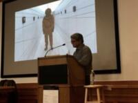  Person speaking at a podium in front of a projected screen showing an image of a person in a perspective hallway.