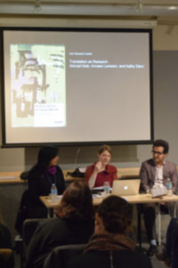 Panel discussion with three speakers in front of an audience. A presentation slide is displayed on a screen.