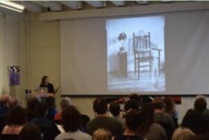 Person presenting to an audience with an image of a wooden chair and a table with a plant projected on a screen.
