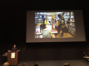 A speaker presents in an auditorium with a large screen showing a library scene.