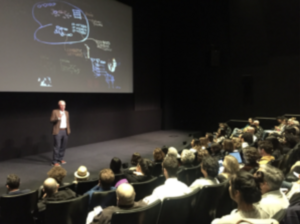 A person gives a presentation in a dimly lit auditorium with a large screen displaying a complex diagram.