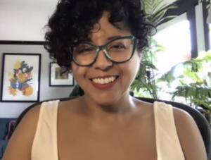 Person with curly hair, green glasses, and white top smiling in a room with houseplants and framed artwork.