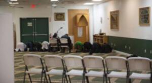 People praying in a hall with chairs in the foreground and framed decorations on the wall.