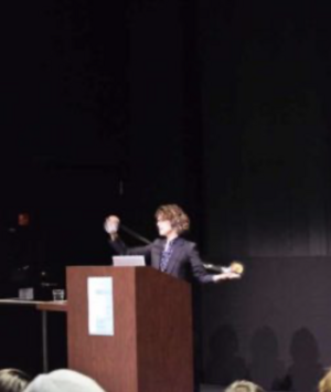 Person speaking at a podium, gesturing with arms in a dark room.