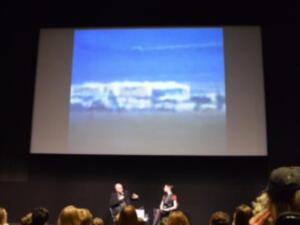 Two people seated on a stage in front of an audience with a large screen displaying a blurry blue and white image above them.