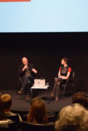  Two people seated on stage engaging in a discussion in front of an audience.