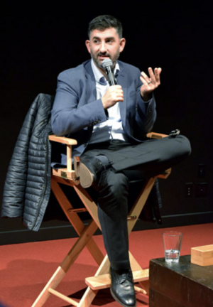 Man seated on a director's chair speaking into a microphone, with a glass of water on a nearby table.