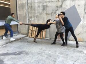 Four people interacting humorously; one person holding another’s boot as they extend horizontally mid-air, supported by two others against a concrete urban backdrop.