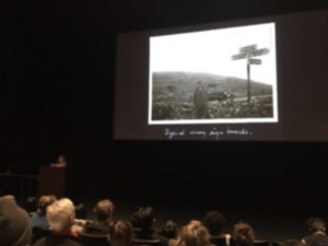 Lecture hall with a speaker at a podium and an audience watching a presentation with a black-and-white photograph on the screen.