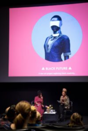 A presentation scene with two women seated on stage in front of a screen displaying a futuristic figure wearing a visor.
