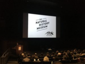 Auditorium with audience facing a screen displaying "THE NATURAL HISTORY MUSEUM" and a dinosaur drawing.