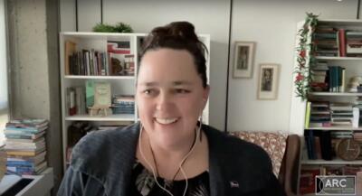  A person smiling with earphones in front of bookshelves filled with books and decorations.