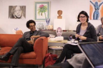 Two people sitting and conversing in an art studio with various artworks and sculptures in the background.